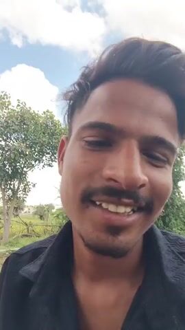 The video features a close-up shot of a young man with dark hair and a mustache, wearing a black collared shirt. He is outdoors, with trees and a cloudy sky visible in the background. The man smiles and speaks to the camera. He says, "I need 5 million dollars. You give me 5 million dollar."