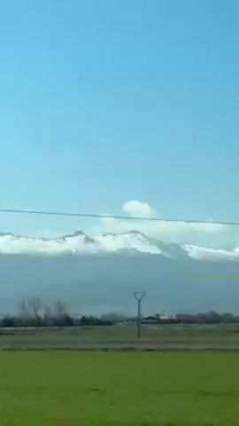The camera moves along a road, viewing a green field, and in the background are mountains with snow on top. An electric utility pole can be seen from the car window.