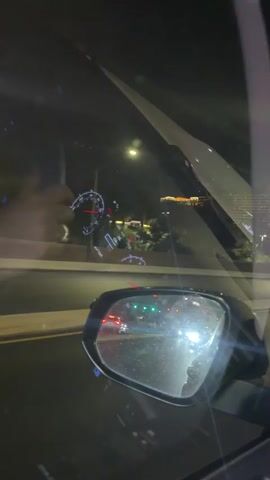 The video shows a view from inside a car driving at night. The camera pans up and to the right, showing the side mirror and a view of the road behind the car, lit by headlights of other vehicles. The driver's hand and ring are visible in the upper left corner of the frame. In the foreground, the car's dashboard and side mirror are visible. Through the windshield, the blurred lights of the Las Vegas strip can be seen. Streetlights illuminate the highway, marked by yellow lines.
