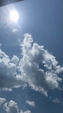 A vertical video shows a bright sun and white clouds against a blue sky. The sun is in the upper left corner and emits bright rays. The clouds are puffy and appear to be moving slowly across the sky. In the lower left corner, a person's head and shoulders are visible from the neck up, wearing a dark shirt. A dark object, possibly part of a structure or an umbrella, is visible in the upper portion of the frame, casting a slight shadow. The video appears to be shot from a low angle, looking upwards.