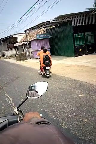 The video begins with a first-person view from a motorcycle, showing a street scene. Two individuals in orange clothing are on a scooter, stopped on the side of the road. One is driving, and the other is a passenger. A small purple kiosk with the sign "ES KACANG MERAH" is behind them. The motorcycle continues, and a dark-colored car drives past in the opposite direction. Further down the road, a yellow truck is visible. The motorcycle then approaches and passes the yellow truck. The road appears to be in a somewhat rural or developing area, with modest buildings and power lines visible. The weather is sunny.