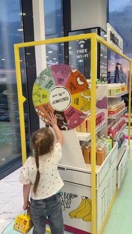 A little girl stands in front of a Dolce Milk promotional display and spins a wheel of flavors. The child is seen from the back, with a long blonde braid and wearing jeans and a white t-shirt with pink hearts. The wheel, framed in yellow, has a variety of flavors listed, including Strawberry, Banana, Tangerine, and Pear. The display is well-stocked with rows of colorful bottled products. It also has cartoon-style drawings of bananas on the bottom. The environment suggests an indoor location, possibly a shopping mall. There is a window behind the display which provides a view of a parking lot outside. A sign advertising "FREE WI-FI" is visible at the top corner of the display.