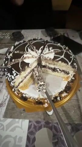 A chocolate cake is shown on a table, partially cut. Someone is holding a piece of cake with a utensil, and another person is seen with cake on their face. The environment appears to be indoors, possibly a kitchen or dining area, with wooden walls and cabinets visible.