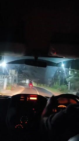 The video shows a first-person view from inside a car at night. The driver's hands are visible on the steering wheel. The dashboard lights are illuminated, with a red screen display in the center and orange lights on the speedometer. Outside the car, the road is visible, illuminated by streetlights. A motorcycle with a red light on the back is in front of the car, moving away. Buildings and trees can be seen on the sides of the road. The car appears to be in motion.