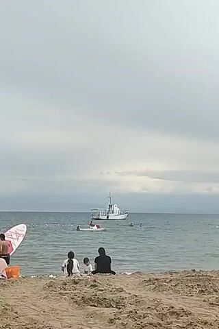A wide shot shows a cloudy day at the beach. In the foreground, three people sit on the sand facing the water. Two children, one wearing a white shirt and the other a red shirt, sit between two adults. One adult wears a black headscarf and the other is not visible. In the middle ground, several people swim in the ocean. A white boat with a red stripe is anchored in the water. In the background, land is visible under a cloudy sky.
