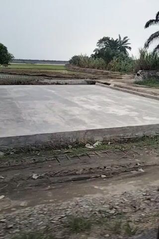 The video, oriented vertically, shows a dirt path next to a building with a corrugated metal roof. Two motorcycles are parked near the building. Beyond the building, there are trees, including palm trees, and a body of water with a gate structure. The path continues past the water, with more vegetation, including what appear to be banana trees, and then past another building with a window. The ground is uneven and appears muddy in places.