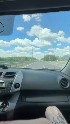 The video is a first-person perspective from inside a car driving on a highway on a sunny day. The dashboard and steering wheel are visible in the foreground. A stuffed toy is hanging from the rearview mirror. The road ahead is clear with some trees and greenery on either side. A gas station with a tall sign is visible in the distance on the right. The sky is blue with scattered white clouds. The camera pans slightly, showing the driver's hands on the steering wheel and the passenger's legs.