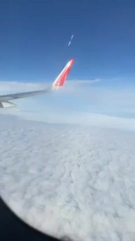 The scene is a view from an airplane window. The sky is bright blue with wisps of white clouds, and the wing of the plane, with a red and white tip, is visible. Below the plane, there's a sea of fluffy white clouds stretching to the horizon. The edges of the window frame are visible.
