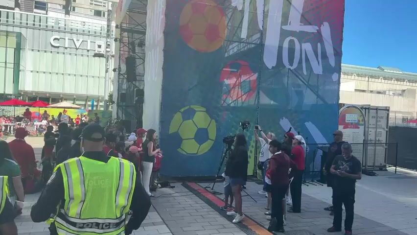A group of people are gathered around a large banner featuring soccer balls. A woman holding a microphone appears to be interviewing a young person. There are several cameras filming the scene. A police officer in a reflective vest is standing in the foreground, facing away from the camera. In the background, there is a building with the word "CIVIC" visible. The ground is paved with bricks. The overall atmosphere seems to be a public event or media coverage.