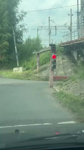 The video shows a red traffic light at a railroad crossing. The traffic light is mounted on a wooden pole, and there is a wooden barrier extending over the road. The railroad tracks are visible in the background, along with overhead power lines. The area is overgrown with grass and trees. The video appears to have been filmed from inside a vehicle.