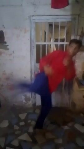 A young person in a red, long-sleeved shirt and blue pants is performing martial arts moves indoors. The person executes kicks and punches, showing some form of training or practice. The background includes a white door with a metal grate, a wall with peeling paint, and a patterned tile floor. The lighting in the room appears dim, adding a slightly gritty feel to the video.