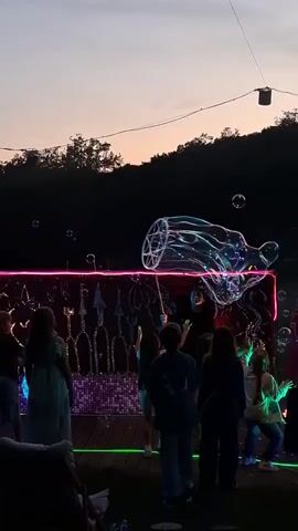 The video shows a group of children watching a bubble show at dusk. They are standing in front of a stage that is decorated with lights. In the background is a line of trees and a pink sky. The bubble artist is making large, elaborate bubbles. The children are pointing and clapping.