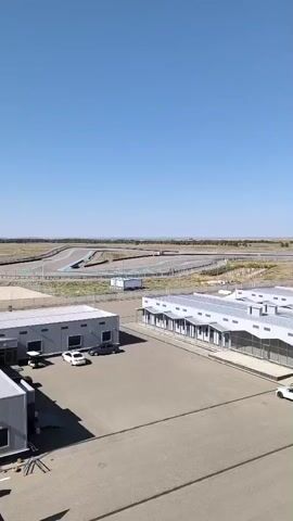 The video opens with a wide shot of an empty, sun-drenched outdoor area, possibly a testing facility or car lot, with a racetrack visible in the background under a clear blue sky. Several modern, grey-roofed buildings are lined up in the foreground. As the camera zooms in and pans down, the scene shifts to focus on a collection of cars parked in a lot between the buildings. A person in dark clothing walks between the parked vehicles. The vehicles appear to be a mix of sedans and SUVs, predominantly in dark colors, with a few lighter colored cars interspersed. A light blue sports coupe is prominently positioned in the center of the group. The camera continues to move, offering a closer view of the cars and the person walking among them. In the final frames, the camera continues to pan and zoom, showcasing more of the parked vehicles.