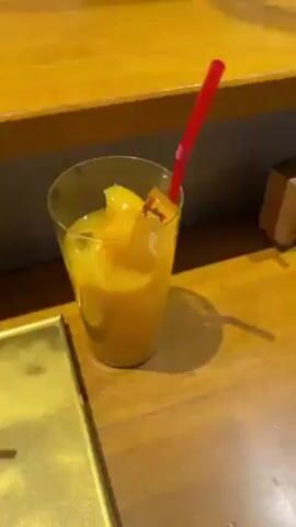 On a wooden countertop, a full glass of yellow drink sits next to a golden tray. Next to the tray is a white bowl containing noodles with a reddish-orange sauce and what appears to be slices of black truffle. A wooden spoon rests in the bowl. The bowl has the word "MARCO" written on the side.