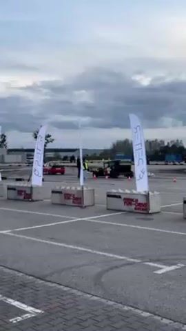 The video shows a black hot rod driving in circles in a large parking lot or driving course. In the background, a red car is also driving. Several people are standing around the driving course, observing. The sky is overcast. In the foreground, there are four gray concrete blocks with the words "MANOL FUN-DRIVE DZIECIOM" printed on them, each block having a white flag with the word "EPETTER" on it.