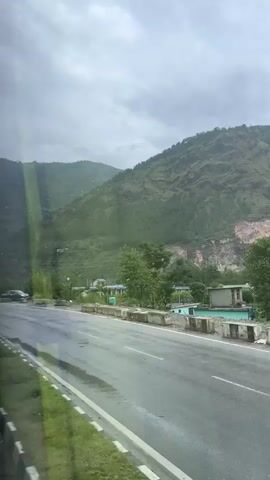 The video shows a cloudy day on a wet road. Mountains covered in green trees rise in the background. A roadside building with multiple stories is visible. A truck, a motorcycle, and a bus are present on the road. Traffic signs indicate a curve in the road.