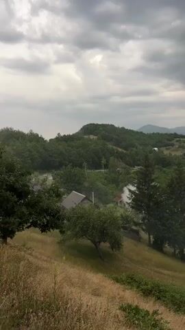 A panoramic shot captures a cloudy, overcast sky over a lush, green mountainous landscape. In the foreground, a grassy slope is dotted with dry vegetation and a few trees, including a lone, gnarled tree. A rustic house with a red-tiled roof and a chimney is visible on the right side of the frame. In the mid-ground, nestled amongst dense trees, are a few scattered houses and structures. Smoke billows from several points in the valley, dissipating into the misty air, suggesting either fires or possibly agricultural burning. The overall atmosphere is serene and rural, with a hint of mystery due to the rising smoke.