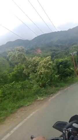 The video captures a scenic view of a mountainous landscape with lush vegetation. A paved road is visible in the foreground, with power lines stretching across the sky. The terrain is characterized by rolling hills covered in trees and foliage. The sky is partly cloudy, with sunlight intermittently breaking through. The overall impression is one of natural beauty and tranquility.