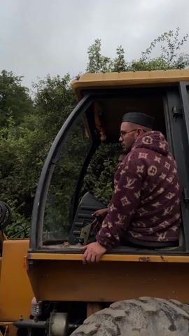The video shows a man sitting in the cab of a yellow construction vehicle. The man is wearing glasses and a dark kufi hat. He is also wearing a patterned fleece jacket. He is looking to the left and appears to be talking on a phone. The construction vehicle has large tires and a boom arm that extends to the left. The background is filled with green trees and an overcast sky. The video is filmed from a slightly elevated angle.