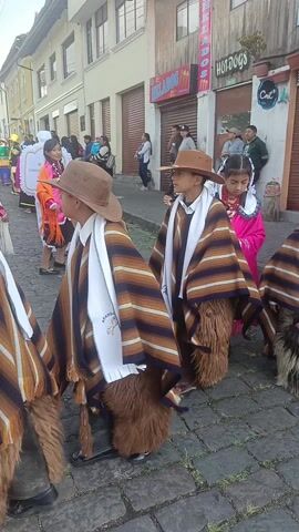 The video shows a group of children participating in what seems to be a street parade or cultural event.  The children are dressed in traditional attire. Some of the boys are wearing brown hats, striped ponchos, white scarves, and furry leggings.  The girls are wearing colorful dresses and shawls.  They are standing on a cobblestone street.  In the background, there are buildings with closed storefronts, advertising "Helados" and "Hotdogs." People are standing on the sidewalks watching the event. A piercing whistling sound, likely from a musical instrument, is heard throughout the video.