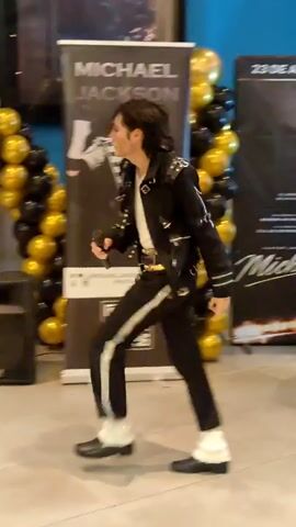 A person dressed as Michael Jackson is dancing and singing in front of a backdrop with balloons and a banner that says "MICHAEL JACKSON". The person is wearing a black jacket with white stripes and accents, black pants with white stripes, white socks, and black shoes. They are also holding a microphone. The backdrop has a QR code and a poster of Michael Jackson. The environment appears to be an indoor event space, possibly a movie theater or shopping mall.