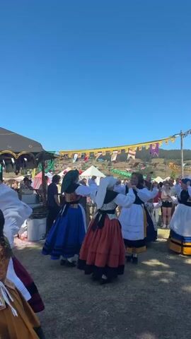 The video shows an outdoor event with several people. In the foreground, a group of women are dancing in traditional folk costumes. They are wearing full skirts, ornate bodices, and head coverings. Some of the skirts are blue and black striped, while others are orange and black. One woman in the foreground is facing away from the camera and her skirt is a mix of yellow, orange, and green. Behind the dancers, there are tents, stalls, and a crowd of onlookers. The sky is clear and blue, suggesting it is a sunny day. The overall atmosphere appears festive.