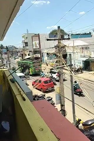 The video captures a busy street scene from a balcony. The street is filled with various vehicles, including cars, motorcycles, and a green bus. There are also a number of people walking around. The buildings lining the street appear to be shops and residences. A prominent feature of the scene is the extensive network of electrical wires and poles that crisscross the sky above the street, creating a tangled web. The sky is blue with scattered clouds, suggesting a sunny day. The perspective from the balcony suggests the viewer is looking down and across the street, with a railing in the foreground.