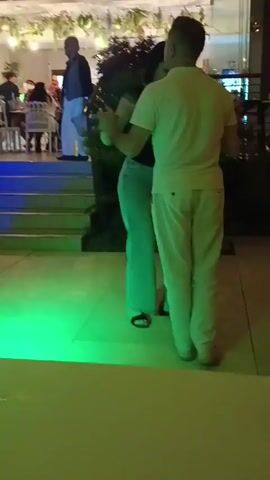 This video, filmed in Roseto degli Abruzzi, Italy, captures a lively evening scene outside a restaurant or bar. The primary focus is a couple dancing and kissing in the foreground, illuminated by blue stage lights and the ambient glow of the establishment. The man is wearing a light-colored t-shirt and pants, while the woman is in a dark top and light blue jeans. Their embrace suggests an intimate and affectionate moment.

In the background, several other people are visible. Some are seated at tables inside the venue, while others stand or walk around. One man in a dark jacket and light pants is standing on the steps leading up to the establishment, facing away from the camera. Another person, with short, light-colored hair and wearing a white t-shirt and black shorts, walks past the dancing couple, momentarily blurring into motion.

The setting appears to be an outdoor patio or walkway. The ground is paved with light-colored tiles. Behind the dancing couple, there are lush green plants. The establishment itself has a modern facade with large windows, and decorative hanging greenery is visible above the entrance. The lighting suggests it is evening or nighttime, with artificial lights casting colorful hues on the scene. The overall atmosphere seems festive and social, with people enjoying themselves. No visible text is present in the video.