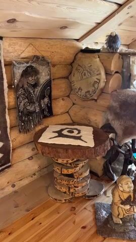The video shows the interior of a rustic-style wooden building. The walls are made of logs, and there are various decorative items throughout the space. A small, round wooden table with a symbol on it sits near the wall. Other items include a carved figure, animal furs, and what appear to be shamanic drums and artwork on the walls. The camera pans upwards to show an upper level with more drums and decorative items displayed along a railing. The room is well-lit by natural light coming through large windows. A man is visible in the background on the lower level, wearing a white t-shirt. He appears to be looking at a display of pottery.