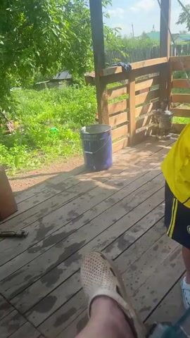 A low-angle POV shot shows a person's foot in a white clog, resting on a wet wooden deck. To the left, a brick lies on the deck, and a black pole extends towards it. In the background, a blue bucket sits near a wooden fence. Green foliage is visible behind the fence. A person wearing a yellow shirt and dark shorts walks into the frame from the right. The camera then shifts to a blurry close-up of skin, possibly an arm or leg, showing reddish marks.