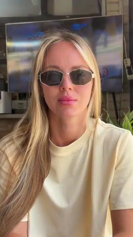 This media file features a medium close-up shot of a young blonde woman wearing a pale yellow t-shirt and gold-rimmed sunglasses. Her hair is long and wavy, with some blonde highlights. She is looking directly at the camera with a neutral expression.

Behind her, a television screen displays a colorful image that appears to be from a sporting event or a movie, with text visible that reads "IGNITION" in large white letters. The setting seems to be outdoors, possibly a cafe or a patio, with some greenery visible on the right side of the frame. The lighting suggests it is daytime. The woman's attire and the relaxed setting hint at a casual, possibly warm-weather occasion. There are no strong indicators of emotion, giving the impression of a posed or candid moment without significant emotional expression. The background is slightly out of focus, drawing attention to the woman.