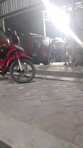A red motorcycle is parked on the left side of the frame. In the background, a red truck is visible, with people around it. Another red vehicle, possibly a tractor or another truck, is parked behind the first truck. The scene is illuminated by bright overhead lights, creating strong shadows. The ground is paved with interlocking bricks.