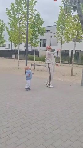 A man in a grey tracksuit with black stripes on the legs is playing basketball with a young child on an outdoor court. The man is bald and has a beard. He is in the process of shooting a red basketball into a hoop. The child, wearing a denim jacket and grey pants, is watching him. The court is paved with grey interlocking bricks. There are trees with green foliage and modern buildings in the background. The sky is overcast.