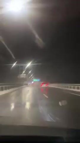 A car travels on a highway at night during winter weather conditions. The roadway is wet and slushy. Streetlights illuminate the scene. Taillights of other cars ahead are visible. The point of view is from inside the vehicle looking through the windshield, which has some water droplets on the glass.