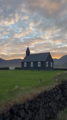 A picturesque static shot captures the iconic black church of Búðir, Iceland, set against a stunning sunset or sunrise sky. The church, with its dark, vertical wooden siding and contrasting white window frames and door, stands prominently in a vibrant green field. A low wall of dark, volcanic rocks forms the foreground, suggesting the rugged natural landscape. In the background, rolling mountains are silhouetted against the horizon, where warm orange, yellow, and soft pink hues peek through a ceiling of scattered clouds. The scene conveys a sense of tranquility and natural beauty, characteristic of Iceland's unique environment. No people or visible text are present in the frame.