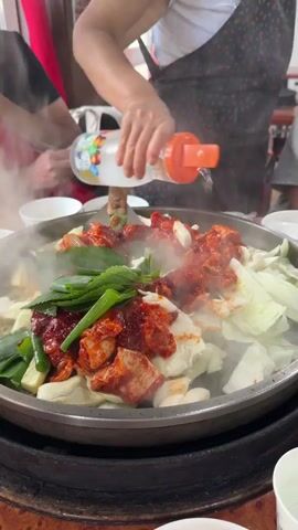 A person in a black apron with a white top is pouring liquid from a clear plastic bottle with an orange cap into a large metal pan filled with food. The pan contains chicken pieces coated in a red sauce, shredded cabbage, and green onions. Steam is rising from the pan, indicating it is hot. Another person in a red apron is visible in the background, holding chopsticks and seemingly eating. The food is being cooked on a circular black stove. The setting appears to be a restaurant, with wooden tables and bowls visible.