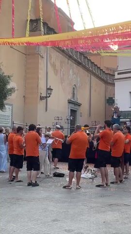 A brass band comprised of approximately ten people, all wearing matching bright orange t-shirts and dark shorts, plays instruments in an outdoor public space. The band is arranged in a semi-circle facing an audience, which is partially visible in the background. The musicians are playing brass instruments, including trumpets and a tuba. The ground in front of the band is covered in scattered petals. Behind the band is a large building with a light-colored facade and a red-tiled roof. Colorful banners are strung across the area above the band. Several audience members are observing the performance, with some standing and others appearing to be sitting. The overall atmosphere suggests a festive outdoor event.