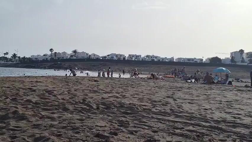 A wide shot of a beach with dark sand shows a group of people scattered across the scene. In the foreground, the sand is undisturbed, showing textures from wind or waves. In the middle ground, closer to the water, a number of people are visible. Several individuals are in the water, while others are standing on the sand, some in small groups. Further to the right, a larger gathering of people are seated on the sand, with one person under a blue umbrella. Behind the beach, a row of white buildings with dark roofs stretches along the coastline, some with palm trees in front of them. The sky is overcast and bright, suggesting a daytime scene. The overall impression is of a moderately populated beach on a clear day.
