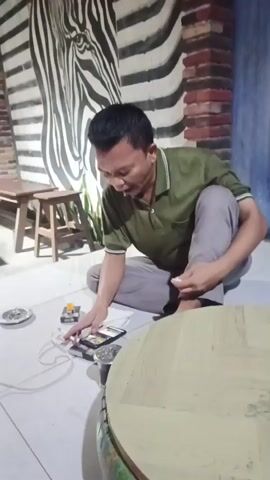 This video appears to capture a candid moment within a cafe or similar establishment, likely in Balongbendo, Indonesia. The primary subject is a man in a green polo shirt and grey pants, crouched on the tiled floor. He is interacting with his smartphone, which is plugged into a power source via a white cable. Several other objects are scattered around him on the floor, including a small ashtray with what looks like a cigarette butt, and a yellow object that might be a lighter or another small electronic device.

In the background, another person is seated at a wooden table, also looking at their phone. The setting features a distinctive wall mural of a zebra's head with black and white stripes, and exposed brickwork. The lighting suggests it could be daytime, possibly afternoon or early evening, given the ambient light and the indoor setting. The atmosphere seems relaxed and ordinary, with both individuals engaged with their personal devices. There is no discernible weather information from the video. The man's facial expression is one of focus as he handles his phone.