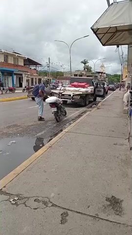 The video captures a street scene. In the foreground, a person is seen interacting with a motorcycle parked on the side of a road. The person appears to be leaning over the motorcycle, possibly adjusting it or retrieving something. Puddles of water are visible on the road surface. In the background, several buildings line the street, some appearing to be shops with awnings. A truck is parked on the right side of the road, loaded with white sacks. Several other motorcycles are parked in front of one of the buildings. Further down the street, a car is parked. Two individuals are walking across the street in the mid-ground, in front of the buildings. The sky is overcast.