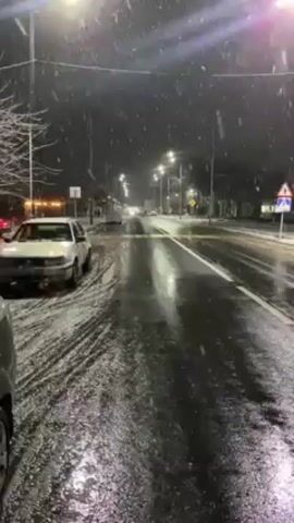 The scene is a nighttime view of a snow-covered street in a residential area. Streetlights illuminate the wet asphalt road, creating bright reflections. Snowflakes are visibly falling. On the left side of the road, a white car is parked at an angle. As the camera pans to the right, a silver car drives past. A building with lit windows is visible across the street. A bus stop shelter with benches is also visible, all covered in snow.