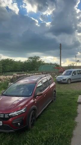 The video shows a rural scene under a cloudy sky with patches of blue. In the foreground, a red Dacia Jogger car is parked on grassy land. In the background, there's a flock of sheep behind a wooden fence. Beside the Dacia, there's a silver van. The camera pans right, revealing a small stone structure, piles of wood, and then focuses on a concrete pipe with objects on top. Beyond the pipe are trees and rolling hills in the distance.