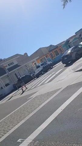 The video shows a pedestrian crossing a street on a sunny day. The street has crosswalks, and there are cars parked on both sides. A man is walking across the crosswalk, and a few cars are visible in the background. The sky is clear and blue. There are buildings on both sides of the street, some with colorful facades. A palm tree is visible in the corner of the frame. The camera angle is slightly angled, and the view is partially obstructed.