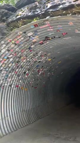 The video begins with a close-up view inside a corrugated metal tunnel, its interior walls covered with numerous colorful stickers. The tunnel curves away into darkness, illuminated by a single overhead light fixture visible in the distance. As the camera pans forward, faint silhouettes of people can be seen walking deeper into the tunnel. The perspective then shifts to a wider shot looking out from the tunnel opening. Beyond the tunnel, a majestic waterfall cascades down a sheer rock face. Lush green vegetation grows around the waterfall and the rocky cliffs. The sunlight catches the spray from the waterfall, creating a misty glow. The lower portion of the video shows the tunnel's entrance from the outside, again with the interior walls adorned with stickers.