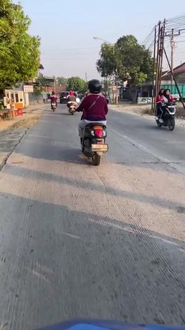 A person wearing a maroon shirt and a black helmet rides a scooter down a paved road. The scooter has a license plate that reads "E 2892 UEH". The person is following a line of other scooters and a car. The road is lined with trees and buildings. The sun is shining, casting long shadows on the road. The video appears to be filmed from another scooter, which is slightly to the right of the center of the frame.