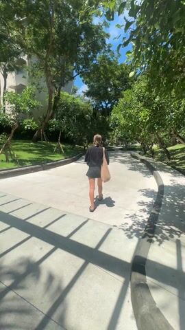 A young woman is walking away from the camera down a concrete path. The path is surrounded by lush green trees and grass on both sides. The woman is wearing a black shirt, dark shorts, and flip-flops, and she has a light brown tote bag slung over her shoulder. The sky above is bright blue with a few scattered clouds. The sun is shining, casting shadows of the trees across the path. In the background, a multi-story building can be seen, partially obscured by the trees.
