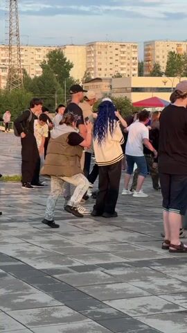 A group of people are gathered outdoors in a paved area. In the foreground, a person wearing a brown puffer vest and patterned pants is facing away from the camera, interacting with another person who has blue hair and is wearing a white top and black pants. To the left of this pair, several individuals are standing and conversing, mostly dressed in dark clothing. In the background, there are tall residential buildings and a metal tower. The overall atmosphere appears to be casual, possibly a public gathering or event.