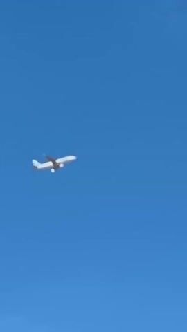 An airplane is flying in a clear blue sky. The airplane is white with a blue tail. It is angled slightly upwards. There are a few wispy white clouds in the distance. The video is brief, showing only a few seconds of the airplane's flight.