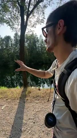 The video pans across an outdoor scene with two men. The first man, wearing sunglasses and a white t-shirt, is in the foreground, gesturing towards a body of water. Behind him, partially visible, is another man wearing a grey t-shirt, sunglasses, and a baseball cap, with a towel draped over his shoulder. The environment appears to be a park or natural area with trees, water, and a pathway. The sun is shining brightly, creating lens flare in some shots. There is a signpost with multiple signs visible near the water. In one brief shot, a person in a black bikini is seen sitting on rocks near the water. The camera movement is fluid, capturing different angles of the scene and the men's interactions.
