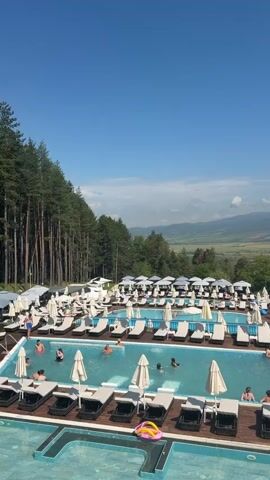 The video captures an overhead shot of a luxurious resort pool area on a bright, sunny day, with clear blue skies and minimal white clouds. A vast landscape of trees and mountains forms the backdrop. The pool area is bustling with activity. Two large pools, one rectangular and one with a unique shape, are visible, surrounded by wooden decking. Numerous white lounge chairs are strategically placed around the pools, some with beige umbrellas providing shade. Several individuals are seen swimming and relaxing in the pools, while others are lounging on the chairs, soaking up the sun. The scene exudes an atmosphere of leisure and tranquility.